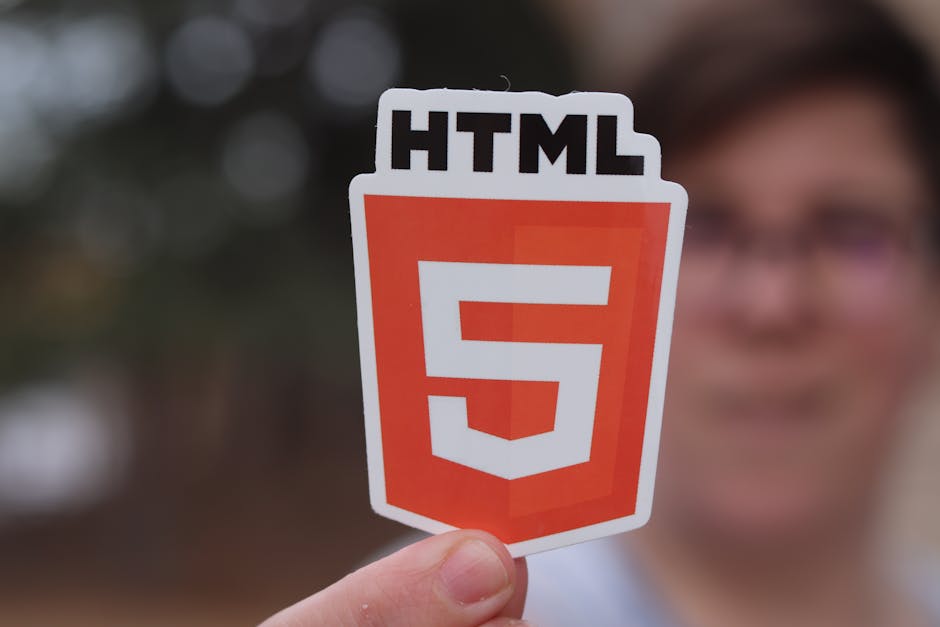 A person holding a rectangular sticker with the HTML5 logo, featuring the word 'HTML' at the top and a large orange shield with a white number '5' in the center. The background is blurred with hints o