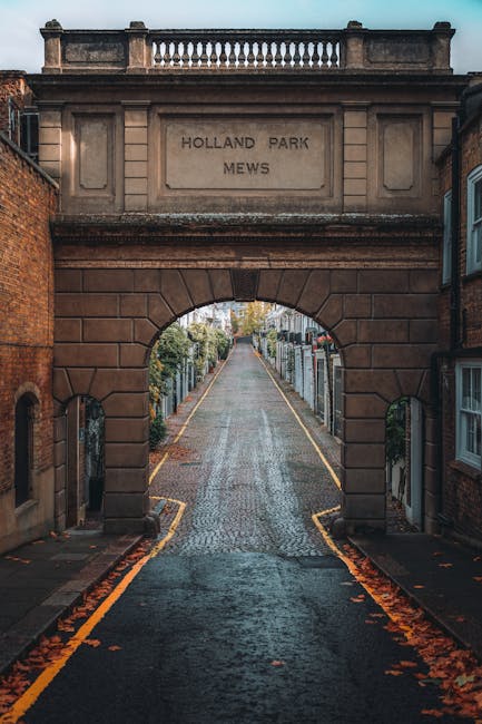 A narrow cobblestone street viewed through a historic stone archway inscribed with 'HOLLAND PARK MEWS.' The street gently slopes upward, bordered by residential gardens and hedges with trimmed bushes 