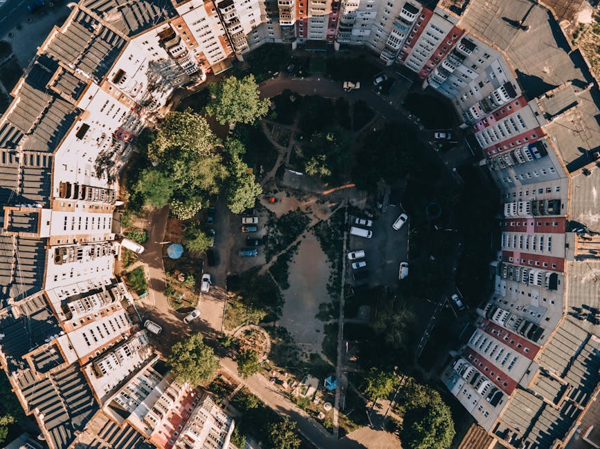 An aerial view of a circular residential courtyard surrounded by multi-story buildings with a variety of colors including white, red, and brown. The courtyard features a central landscaped area with s