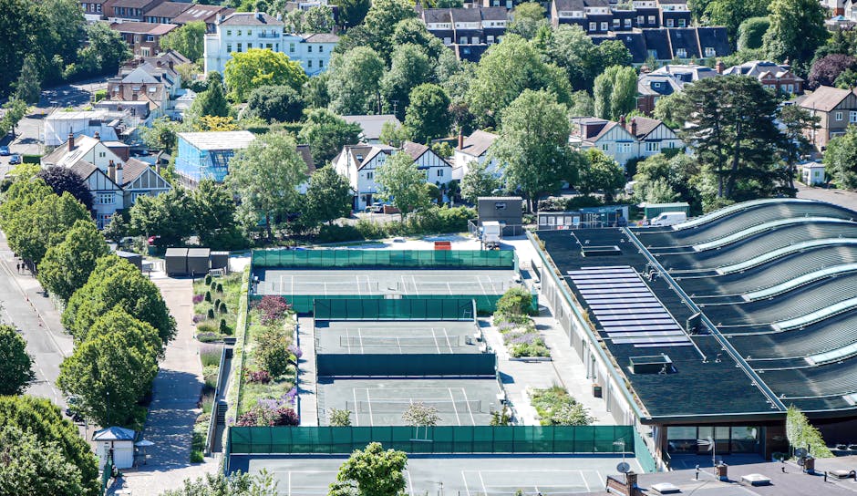 An aerial view of a circular residential courtyard surrounded by multi-story buildings with a variety of colors including white, red, and brown. The courtyard features a central landscaped area with s
